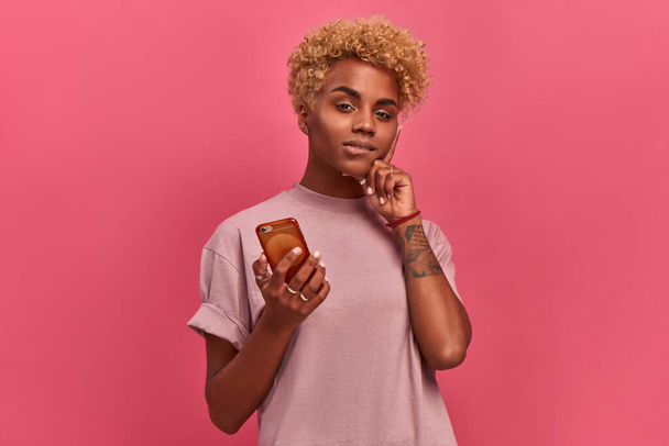 Pensive intrigued pretty african american hipster female with blond curly hairdo holding a smartphone in her hands and chatting in social networks. Targeted advertising concept. Place for text - Foto, Imagem