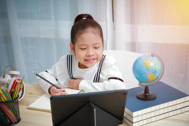 Bambini asiatici che imparano online a casa. Istruzione a distanza per bambini durante il Coronavirus. Scrive risposte e sorride felicemente comunicare con l'insegnante tramite videochiamata
. - Foto, immagini