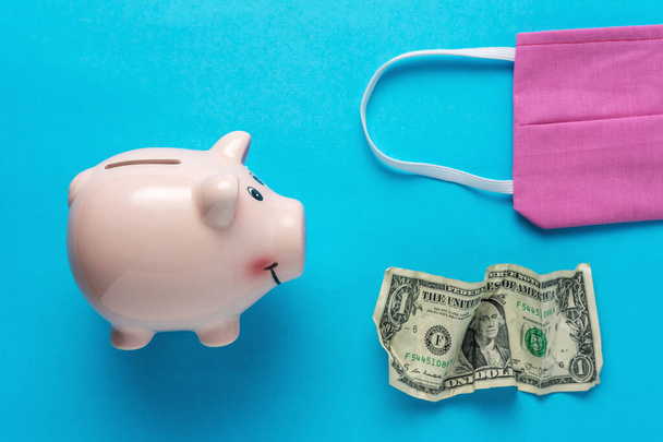 Piggy bank with a medical face mask and crumpled dollar bancnote. Minimal Coronavirus outbreak. Saving money concept. - Fotó, kép