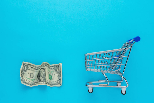 Crumpled dollar bancnote with a shopping cart on blue background. Minimal Coronavirus outbreak. Saving money concept. - Foto, Bild