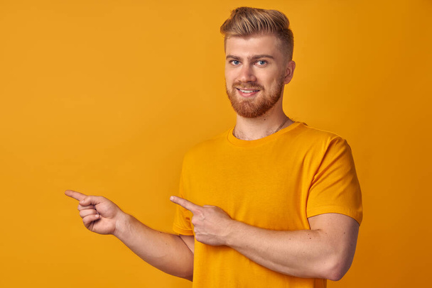 Cheerful young man with beard shows left corner with both index fingers, smiles with approval, suggest going there, on yellow wall. Concept of advertisement of auto repair shop of household appliances - Foto, afbeelding
