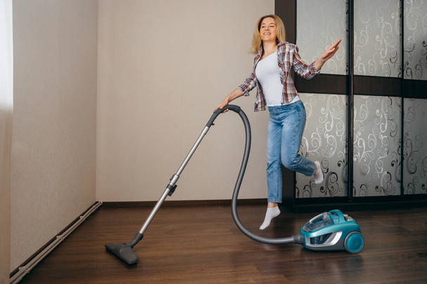 Happy young woman hoovering  laminate at bedroom and jumping.  Funny housekeeping concept. - Foto, afbeelding