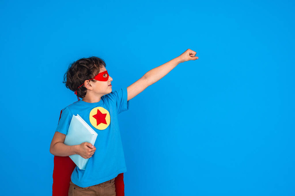 cheerful child dressed in superhero costume holds book and stretches out his hand. little boy laughs and enjoys learning at school. Posing on pink background in Studio Forward to knowledge. - Foto, imagen