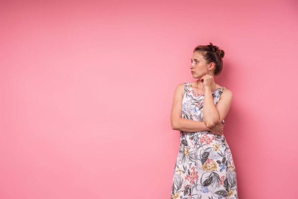 An attractive young woman in long dress with floral print, stands with her hands clasped at her waist and looks thoughtfully to side against a pink background. Concept of waiting space for copying - Fotó, kép