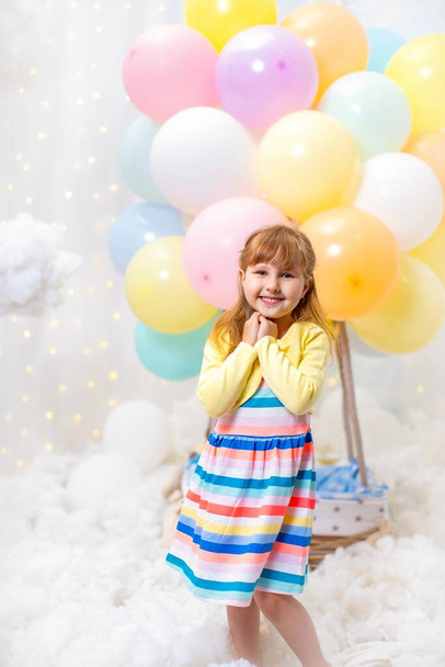 Hayalperest küçük kız dekoratif balon sepetiyle arka planda bulutların üzerinde duruyor. Çocuk çocuk odasında oyun oynuyor. Çocukluk konsepti, doğum günü, tatil süslemeleri. Çocukluk rüyası. - Fotoğraf, Görsel