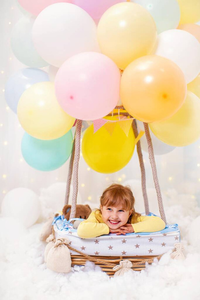 smiling little girl is sitting in basket decorative balloon with her hands under her head. child is playing in children's room. concept childhood, birthday, holiday decorations. Children's dreams. - Photo, Image
