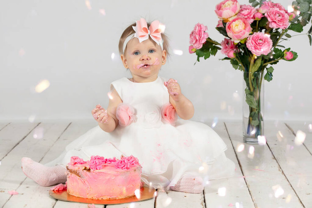 One year old child with a birthday cake. Funny little girl smeared with cream on her birthday. Funny child on holiday. Cute baby girl with pink pastries is sitting on the floor. - Photo, Image
