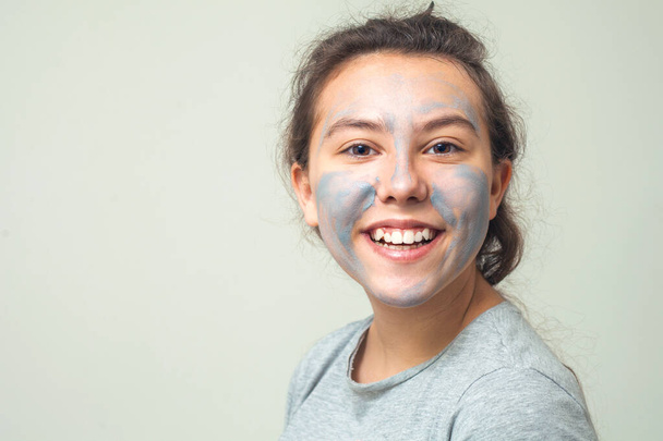 Cute and happy girl smears face with cosmetic clay or mud and smiles. Cosmetic mask, face scrub. The concept of health and beauty. - Photo, Image