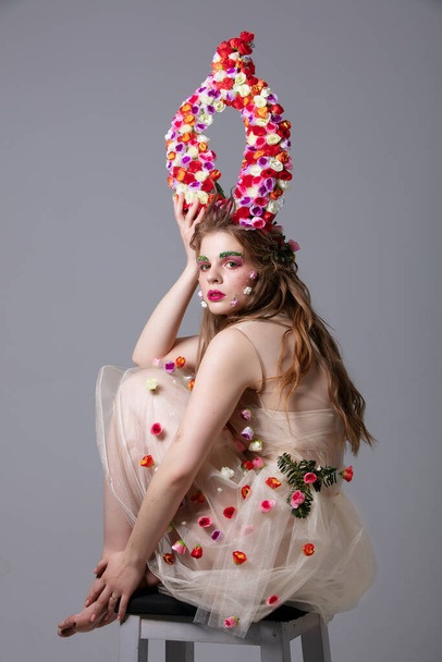 Beautiful girl in a fabulous look with flower horns sits on a chair. Maleficent. Spring or summer beauty. Woman in the image of a flower. - Fotografie, Obrázek