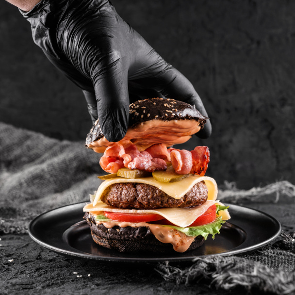 Σεφ χέρι σε μαύρο γάντι μαγείρεμα το μαύρο burger με beaf μπριζόλα, μαρούλι, ντομάτες, φέτα τυρί, ζαμπόν, παστράμι και σάλτσα σε μαύρο φόντο σχιστόλιθο, κοντινό πλάνο - Φωτογραφία, εικόνα