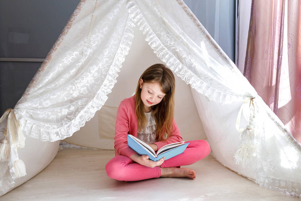 A little girl reads a book at home, sitting on the floor. Reading books in the summer. The child looks at the book with a fascinating smile. - Zdjęcie, obraz