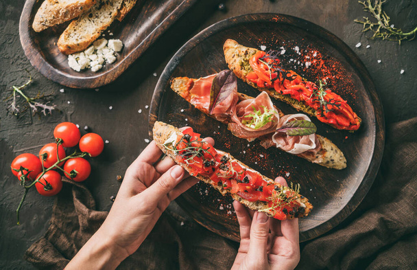 Hands holding a bruschetta with prosciutto, tomatoes, parmesan cheese, sauce pesto, grilled pepper on ciabatta bread on wooden background. Sandwiches set of wine, top view - Photo, image