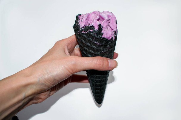 Helado rosa en cuerno negro en la mano de las mujeres aislado sobre un fondo blanco. Vista superior macro cono de helado de carbón con bola de leche suave violeta
. - Foto, Imagen