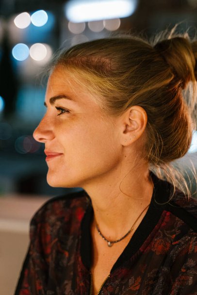 Pionowe zdjęcie portretu młodej blondynki z włosami związanymi na tarasie w nocy ze światłami miejskimi w tle - Zdjęcie, obraz