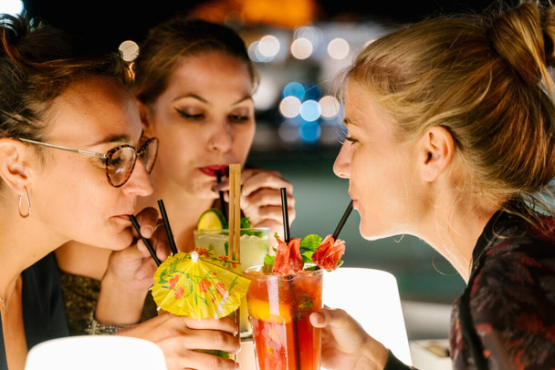 Νεαρές γυναίκες πίνουν κοκτέιλ με καλαμάκι μαζί σε μια βεράντα τη νύχτα με τα θολά φώτα της πόλης στο παρασκήνιο - Φωτογραφία, εικόνα