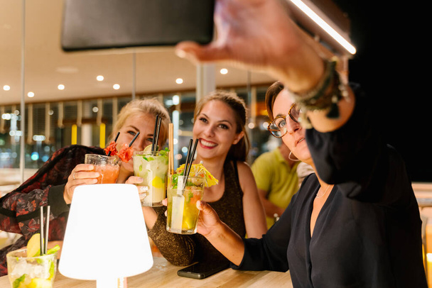 Foto frontale di tre donne che fanno un selfie mentre sono sedute su una terrazza a bere cocktail di notte - Foto, immagini