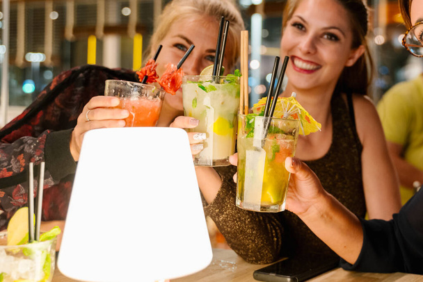 Foco seletivo nos copos de coquetel de três mulheres fazendo uma selfie enquanto estão sentadas em um terraço bebendo coquetéis à noite - Foto, Imagem