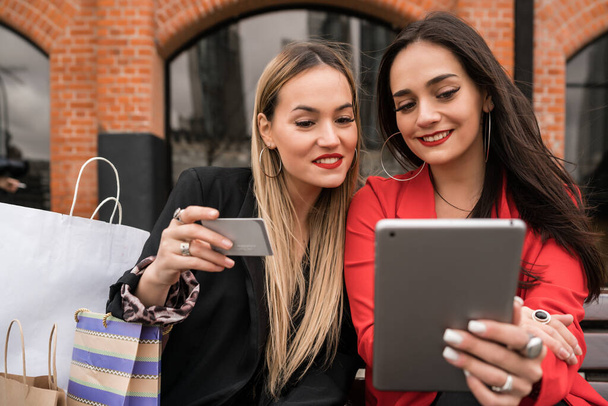 Portrait of two young friends shopping online with credit card and digital tablet while sitting outdoors. Friendship and lifestyle concept. - Photo, Image