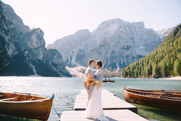 花嫁と新郎は、イタリアのラーゴ・ディ・ブライス(Lago di Braies)の木製ボートドックに沿って歩きます。ヨーロッパでの結婚式、ブライス湖で。新婚旅行、キス、岩の山の背景に抱擁歩く. - 写真・画像