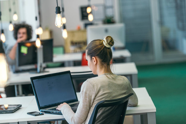 Mulher de negócios loira trabalhando em seu PC enquanto sentada em um escritório moderno lotado. - Foto, Imagem