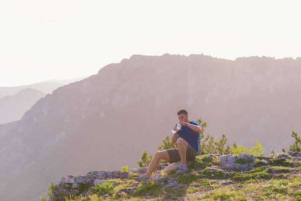 Atleta guapo sentado en un pico rocoso mientras mira la impresionante línea de montaña y un hermoso lago mientras usa una camisa azul y pantalones cortos grises. - Foto, Imagen