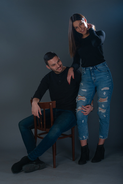 Hombre guapo sentado en una silla y hermosa mujer de pie junto a él sobre fondo oscuro en un estudio
 - Foto, imagen