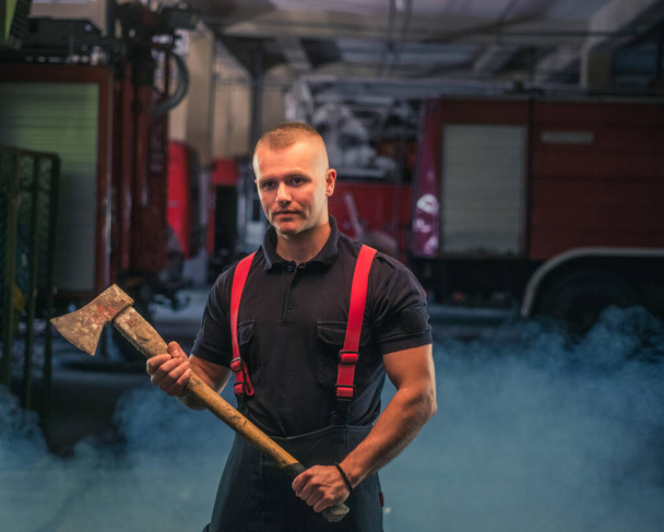 Portrait de jeune pompier tenant une hache dans le garage du service d'incendie - Photo, image