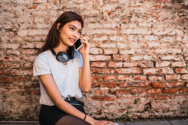Portrait of young latin woman talking on the phone while sitting outdoors against brick wall. Urban concept. - Foto, immagini