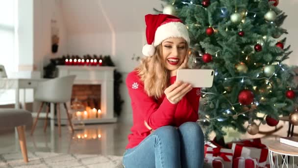 Beautiful young woman in Santa hat and with mobile phone near Christmas tree at home - Кадры, видео