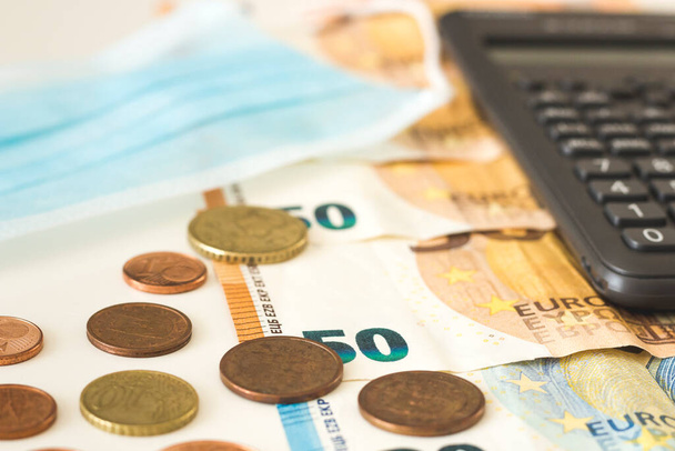 Monedas y billetes en euros de bajo valor junto a una calculadora y una máscara facial. Conceptos, crisis financiera, Covid19
 - Foto, imagen