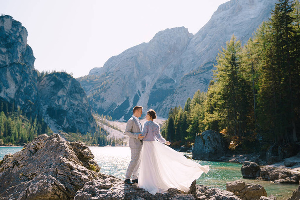La novia y el novio están de pie sobre piedras con vistas al Lago di Braies en Italia. Boda de destino en Europa, en el lago Braies. Amantes recién casados caminan contra el telón de fondo de la naturaleza increíble
. - Foto, imagen