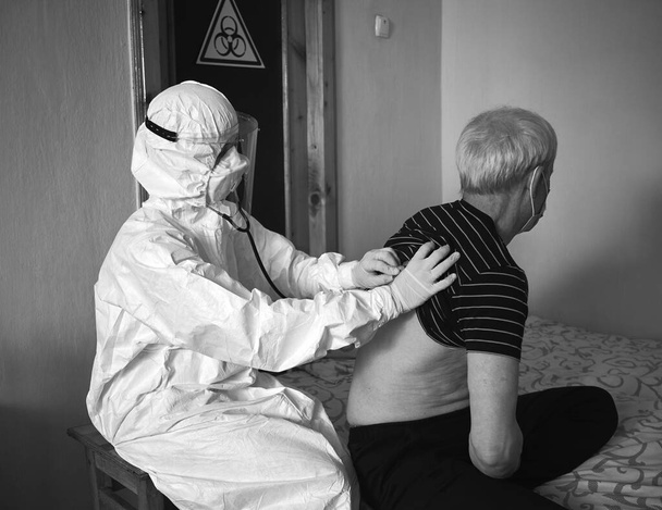 Zwart-wit momentopname van een doktersexamen bij de patiënt thuis tijdens quarantaine 2020. Dokter in beschermend pak en handschoenen controleren geïnfecteerde oude mannelijke patiënt. Biologisch gevarenbord hangt aan de deur - Foto, afbeelding