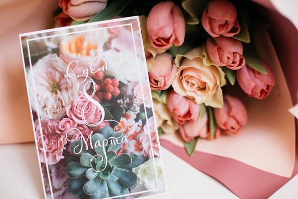 egy csokor friss rózsaszín és őszibarack rózsa papírcsomagolásban, üdvözlőkártyával a fehér asztalon, ajándékként az ünnepre - Fotó, kép