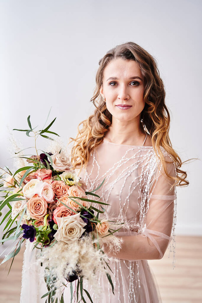 The bride in a dress holds beautiful blooming flowers and looks at the camera isolated on a light background - Photo, Image