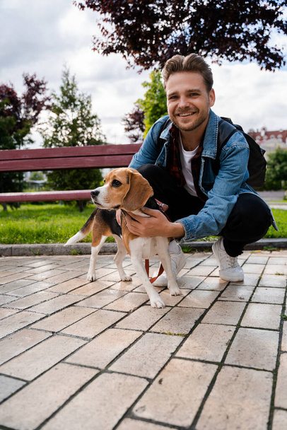 Pystysuora kuva, onnellinen mies hymyilee ja silittää koiraansa puistossa. Ihmiset ja lemmikit, huolehtiva ja hellyyttä lemmikki. Katselen kameraa. Koira ja omistaja kävelevät yhdessä parhaat ystävät
. - Valokuva, kuva