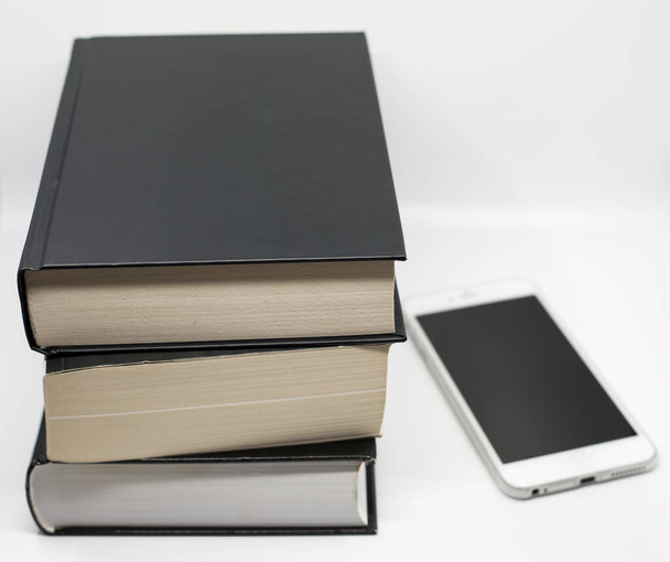Geleneksel kitap mı, kitap mı? Bir yığın kitap, üst üste siyah kapaklı üç kalın kitap beyaz arka planda beyaz bir cep telefonunun yanında. - Fotoğraf, Görsel