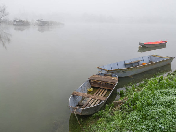 Aangemeerd oude roestige vissersroeiboten aan de waterkant van de rustige rivier en vissershuisjes in de voorkant van rivier eiland diep in de mist. Gloomy herfst landschap verpakt in een dikke mist. - Foto, afbeelding