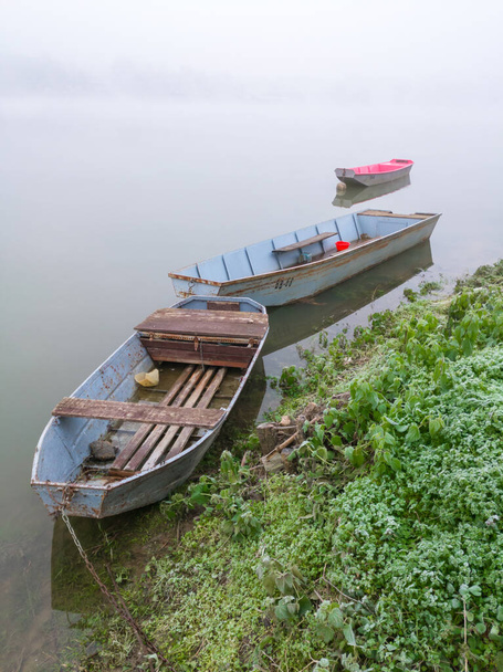 Barche da pesca ormeggiate lungo la riva del fiume nella nebbia profonda. Autunno cupo scena nebbiosa di navi e fiume avvolto in un velo di nebbia
. - Foto, immagini