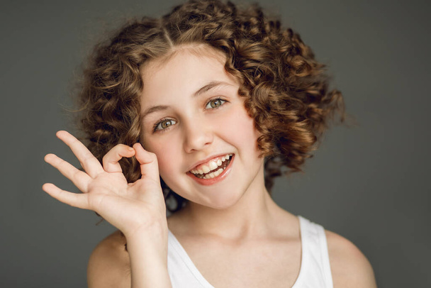 Close-up portrait. A teenager girl with curly hair shows a symbol all is well on her fingers. OK. Gray background. Emotions joy. Delight, Beautiful smile, even teeth. - Photo, Image
