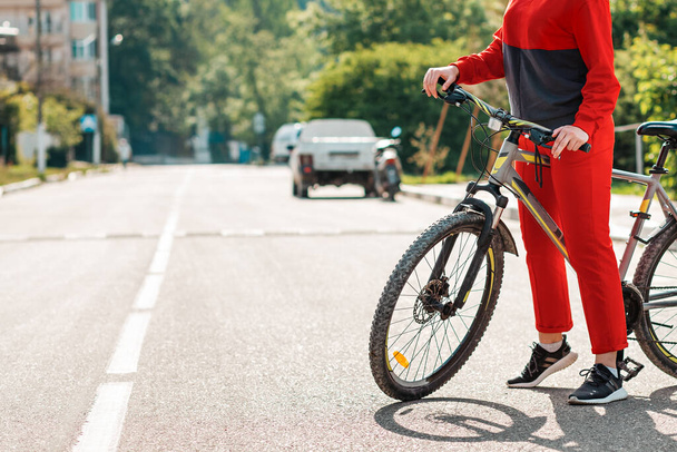 Ciclismo. Una mujer en ropa deportiva roja está de pie con una bicicleta en la carretera. Primer plano de las piernas. Copiar espacio. Las normas de la carretera y el transporte respetuoso con el medio ambiente
. - Foto, imagen