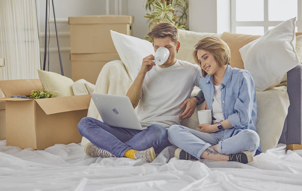 Portrait of happy couple looking at laptop computer together sitting in new house, surrounded with boxes - 写真・画像
