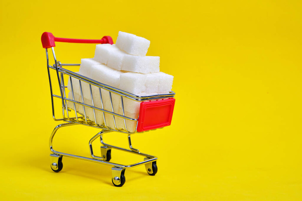 Shopping cart is filled with refined sugar cubes on a yellow background - Photo, image