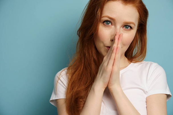 A red-haired girl in a white T-shirt stands on a blue background, pressing her palms to her face. - Foto, Bild