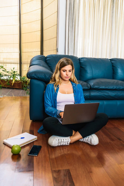Φωτογραφία αρχείου μιας καυκάσιας γυναίκας που κάθεται στο πάτωμα μπροστά από τον μπλε καναπέ της και πληκτρολογεί στο laptop της. - Φωτογραφία, εικόνα