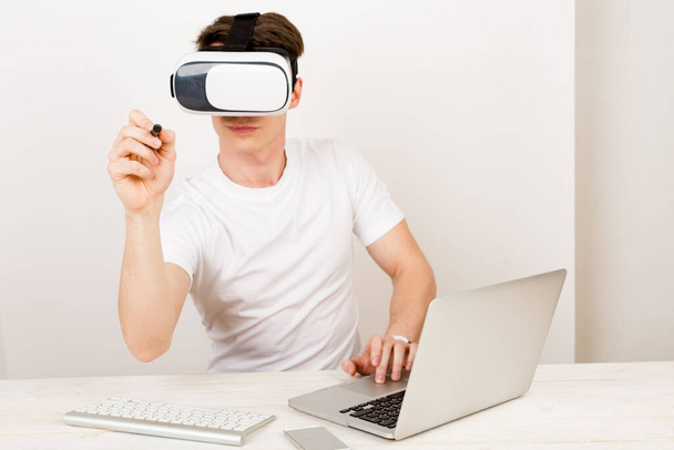 Young man microbiologist using virtual reality headset interacts with augmented things orienting in three dimensional space while sitting on his desk with keyboard and laptop on white background.  - Foto, imagen