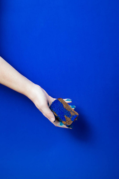 Concept idea: hand holds slice of dark bread covered by blue liquid paint on blue background - Photo, Image