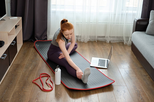 The girl performs a stretching exercise sitting at home in sportsclothes in front of a laptop.  - Foto, Imagem