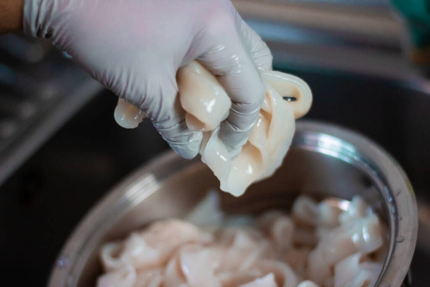 Henkilö puhdistaa mustekalaa lavuaarissa vedellä ja hanskoilla. Tätä mustekalaa käytetään ruoanlaittoon. Kalmarirenkaat ovat metallisäiliössä.
. - Valokuva, kuva