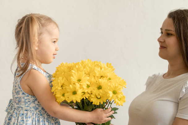 motherhood, mothers day, birthday, childhood, family concept - close-up little girl in blue colorful dress congratulate and give young mother bright bouquet of yellow daisy flowers.on white background - 写真・画像