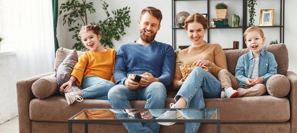 Full body of cheerful young parents in casual clothes sitting on soft big couch with children and watching tv while resting together during weeken - Фото, изображение
